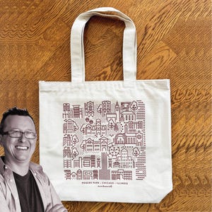 May include: Cream-colored tote bag featuring a detailed, stylized illustration of buildings, trees, and architectural elements. The design is in a dark reddish-brown color. Text at the bottom reads "ROGERS PARK - CHICAGO - ILLINOIS" and "munikeenie". The bag has two handles.