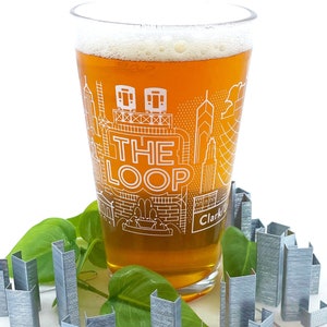 May include: A clear glass beer mug with a white outline of the Chicago skyline and the text "THE LOOP" and "Clark/Lake". The mug is filled with amber beer and has a white foam head. The mug is sitting on a white surface with green leaves and silver staples.