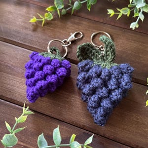 May include: Two crochet grape keychains. One is a deep purple, the other is a dark blue. Both have green leaves and silver key rings. The keychains are on a wooden surface with green foliage.