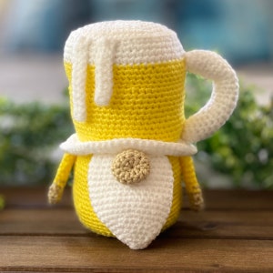 May include: A crocheted gnome shaped like a beer stein. The gnome is yellow and white with a white beard and a brown button nose.