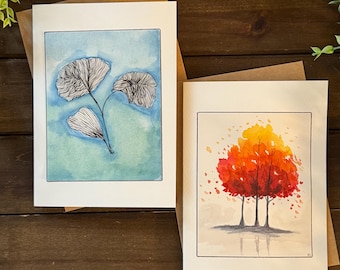 Può includere: Due stampe artistiche ad acquerello su carta bianca, ciascuna con una busta marrone. Una presenta uno sfondo blu con un disegno di foglie di ginkgo in bianco e nero. L'altra raffigura un albero con foglie rosse e arancioni.