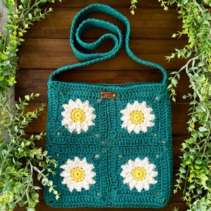 May include: Teal crochet shoulder bag featuring four daisy flower appliqués with white petals and yellow centers. The bag has a long strap and a small leather tag. The bag is handmade and perfect for carrying essentials.