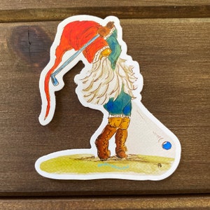 May include: A sticker of a cartoon gnome wearing a red hat and blue pants, swinging a golf club. The gnome is on a green golf course and a blue golf ball is flying through the air.