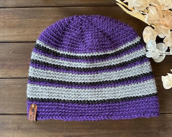 May include: A handmade purple beanie with horizontal stripes in gray and black. The hat is made of a knitted material and has a small brown tag attached to the brim. The beanie is displayed on a wooden surface.