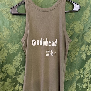 May include: An olive green ribbed tank top featuring the white printed text Radiohead and Pablo Honey on the front. The garment is displayed on a black plastic hanger, showcasing a sleeveless design with a high neckline and a textured fabric.