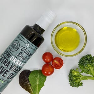 May include: A bottle of raw organic extra virgin olive oil with a label that reads "Village Batch". The image also shows a small bowl of olive oil, cherry tomatoes, and broccoli florets. The bottle contains 16.9 fl oz.