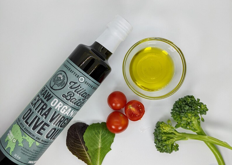 May include: A bottle of raw organic extra virgin olive oil with a label that reads "Village Batch". The image also shows a small bowl of olive oil, cherry tomatoes, and broccoli florets. The bottle contains 16.9 fl oz.