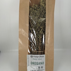 May include: A brown paper bag with a clear window displaying dried oregano. The label reads "Village Batch Organic Greek Oregano." The bag is marked "Made in Greece" and contains 50 grams of the herb.