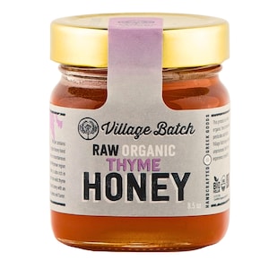 May include: A jar of Village Batch Raw Organic Thyme Honey. The glass jar has a gold lid and a label with the text "RAW ORGANIC THYME HONEY". The jar contains 8.5 oz of honey.