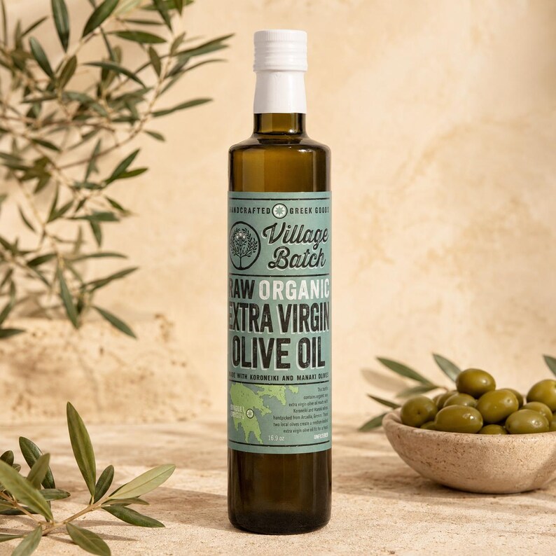 K&ouml;nnte beinhalten: Eine hohe, klare Glasflasche gef&uuml;llt mit dunkelgoldenem Oliven&ouml;l, mit wei&szlig;em Verschluss und hellblauem Etikett. Das Etikett tr&auml;gt die Aufschrift Village Batch Raw Organic Extra Virgin Olive Oil, 500 ml, sowie eine Grafik und Text.