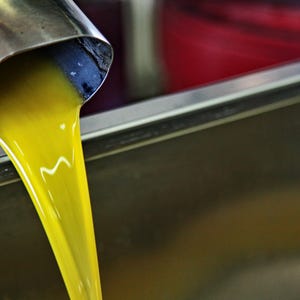 May include: A vibrant, golden-yellow liquid flows steadily from a cylindrical stainless steel spout into a large, metallic basin. The viscous substance catches the light, creating a smooth, continuous stream against the reflective metal surface.