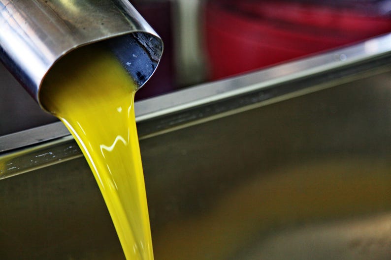 May include: A vibrant, golden-yellow liquid flows steadily from a cylindrical stainless steel spout into a large, metallic basin. The viscous substance catches the light, creating a smooth, continuous stream against the reflective metal surface.