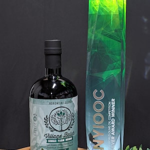 May include: A dark glass bottle of Village Batch single farm origin raw organic extra virgin olive oil with a light green label featuring a tree illustration and the text Koroneiki Olive, next to a tall, faceted green crystal award trophy.