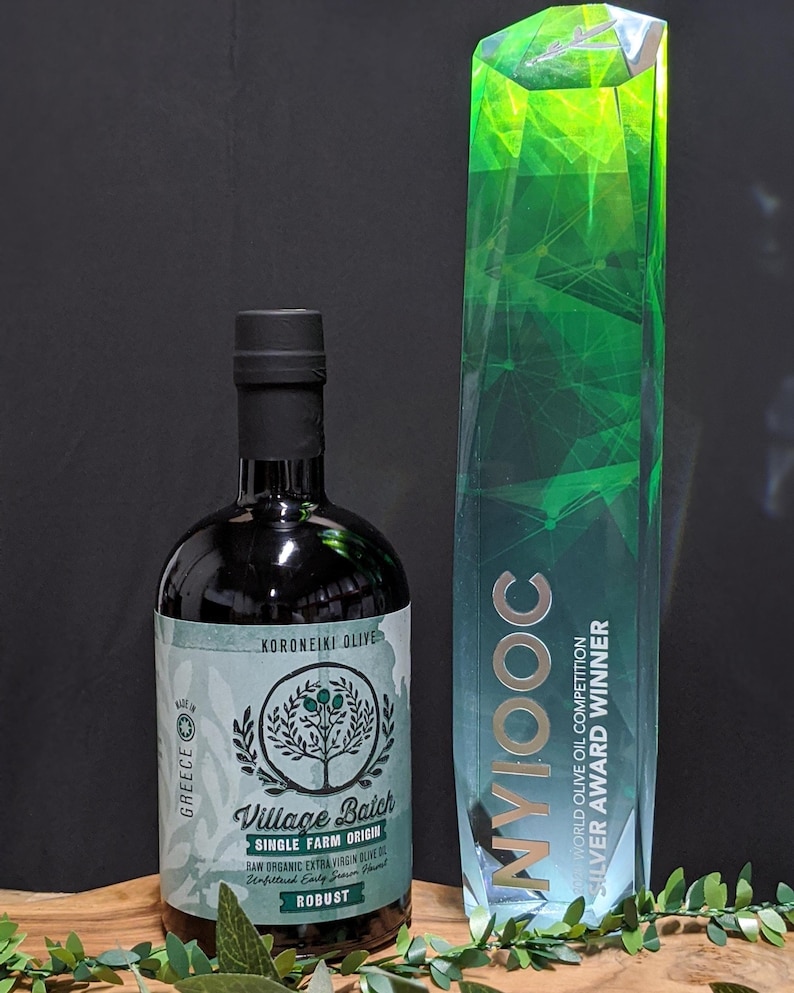 May include: A dark glass bottle of Village Batch single farm origin raw organic extra virgin olive oil with a light green label featuring a tree illustration and the text Koroneiki Olive, next to a tall, faceted green crystal award trophy.