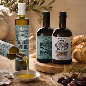 K&ouml;nnte beinhalten: Drei Glasflaschen mit Village Batch rohem Bio-Oliven&ouml;l extra vergine mit Etiketten, die Baumillustrationen zeigen. Eine Flasche gie&szlig;t goldenes &Ouml;l in eine kleine Keramikschale. Die Etiketten spezifizieren die Olivensorten Koroneiki und Manaki.
