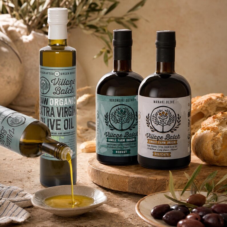 K&ouml;nnte beinhalten: Drei Glasflaschen mit Village Batch rohem Bio-Oliven&ouml;l extra vergine mit Etiketten, die Baumillustrationen zeigen. Eine Flasche gie&szlig;t goldenes &Ouml;l in eine kleine Keramikschale. Die Etiketten spezifizieren die Olivensorten Koroneiki und Manaki.