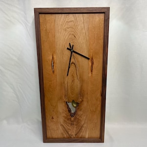 May include: A wooden wall clock with a cherry wood face and a walnut frame. The clock has a unique knot in the wood that is visible through the face. The clock hands are black.