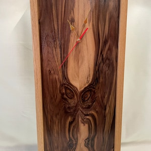 May include: A wooden clock with a natural wood frame and a dark wood face. The clock face has a unique wood grain pattern and gold-colored hands.