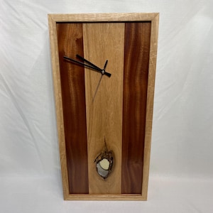 May include: A wooden clock with a natural wood frame and a face with black hands. The clock face is made of alternating strips of light and dark wood, with a knot hole in the center.