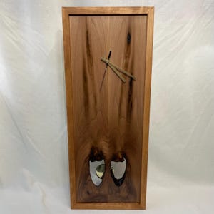 May include: A rectangular wooden wall clock with a natural wood grain finish. The clock features a light brown frame, a dark brown face, and gold-colored clock hands. Two mirrored, oval-shaped insets are near the bottom of the clock.