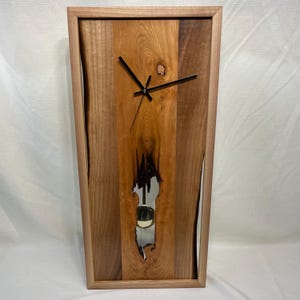 May include: A rectangular wooden clock with a natural wood grain finish. The clock face features black hands and a pendulum visible through a mirrored cutout. The clock is framed in a light-colored wood.