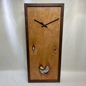 May include: A rectangular wooden wall clock with a light brown face and dark brown frame. The clock hands are black. The face has a unique wood grain pattern with two knots and a heart-shaped cutout near the bottom.