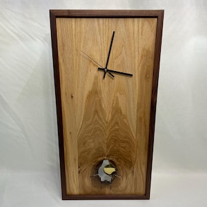 May include: A rectangular wooden wall clock with a light brown wood grain center and a dark brown frame. The clock hands are black. A circular opening near the bottom reveals a gold-colored accent. The clock measures approximately 12 inches wide and 24 inches tall.