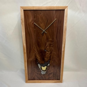 May include: A rectangular wooden wall clock with a dark brown wood grain center and a lighter wood frame. The clock hands are gold-colored. The wood grain has a unique pattern with a lighter-colored section near the bottom.