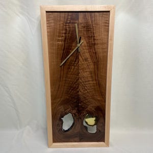 May include: A wooden wall clock with a light wood frame and a dark wood face. The clock has a gold-colored hour and minute hand. The clock face has a unique design with two holes in the wood.