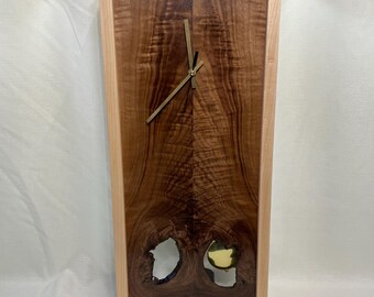 May include: A wooden wall clock with a light wood frame and a dark wood face. The clock has a gold-colored hour and minute hand. The clock face has a unique design with two holes in the wood.