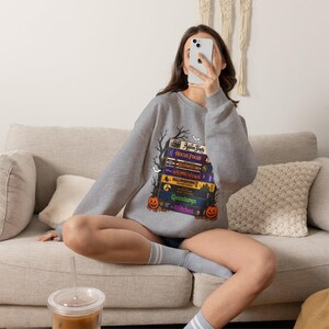 Puede incluir: Una sudadera gris jaspeado de cuello redondo con un estampado gráfico de una pila de cintas VHS vintage con títulos de películas de Halloween como Hocus Pocus, Halloweentown y Goosebumps, flanqueada por siluetas de árboles desnudos y calabazas.