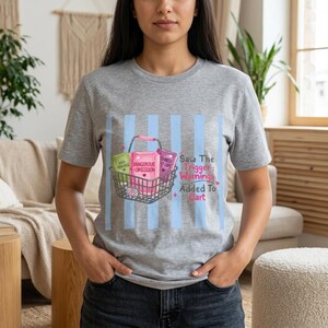 May include: A heather gray t-shirt featuring a graphic of a shopping basket filled with three books, one labeled Dangerous Obsession, alongside the text Saw The Trigger Warnings Added To Cart, set against a background of vertical light blue stripes.