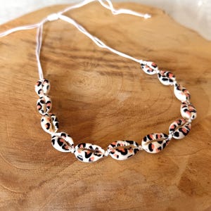 Peut inclure: Collier composé de coquillages cowrie ornés d'un motif léopard noir, rose et orange, enfilés sur un cordon blanc. Les coquillages sont disposés pour former un collier. L'article est présenté sur une surface en bois.