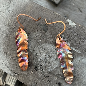 May include: A pair of copper earrings shaped like feathers with a iridescent finish. The earrings are made of hammered copper and have a unique, organic look.