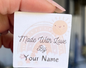 Puede incluir: Etiqueta de tela blanca con un arcoíris, un sol y el texto "Made With Love By Your Name".