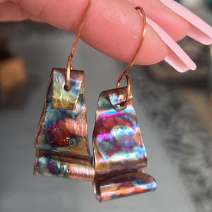May include: A pair of iridescent copper earrings with a wavy, folded design. The earrings are attached to copper hooks.