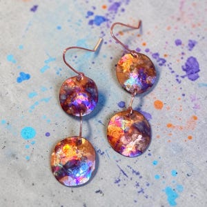 May include: A pair of dangle earrings featuring two circular copper discs each, suspended from thin metal hooks. The discs display a heat-treated finish with a vibrant, iridescent patina showing mottled shades of purple, blue, orange, and gold.