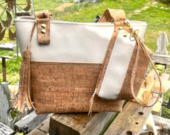 May include: A white and cork tote bag with a smaller matching pouch. The tote bag has a long tassel and the pouch has a gold zipper pull.