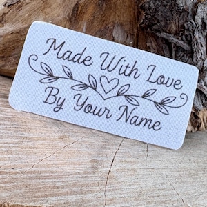 May include: A rectangular, light blue label with the text "Made With Love By Your Name" in black script. The text is framed by a decorative floral and heart design. The label is placed on a rough, wooden surface.