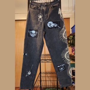 Op de afbeelding: Zwarte denim jeans met verschillende patches en ontwerpen, waaronder een spin, een vogel, een schedel en een skelet. De jeans heeft een wit skeletontwerp op het rechterbeen.