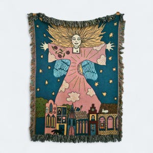 May include: A woven throw blanket with a colourful, whimsical design. The blanket shows a figure with outstretched arms, surrounded by stars, clouds, and a cityscape. The colour palette includes pink, blue, yellow, and green, with fringed edges.
