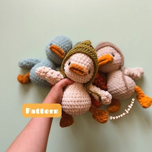 May include: Three crocheted duck plushies in different colors. The duck in the center is white with a green hooded head and orange feet. The duck on the left is blue with a blue hooded head and orange feet. The duck on the right is beige with a brown hooded head and orange feet. The text "Pattern" is visible in the image.