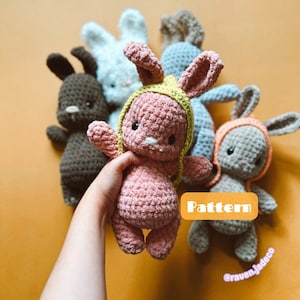 May include: A group of five crocheted bunny plushies in various colors, including pink, brown, white, blue, and gray. The pink bunny is being held in a hand and has a yellow crocheted headband. The text "Pattern" is visible in the image.