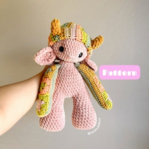 Peut inclure: Un animal en peluche crocheté rose avec un chapeau rayé multicolore. L'animal a un petit corps rond avec deux petits bras et deux petites jambes. L'animal a une petite tête ronde avec deux petites oreilles et deux petits yeux. L'animal tient un panneau qui dit "Pattern".