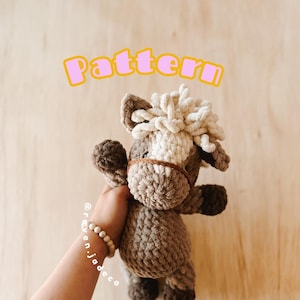 Puede incluir: Un juguete de caballo de crochet marrón y blanco con una melena esponjosa. El juguete está siendo sostenido por una mano con una pulsera de cuentas de madera. El texto "Pattern" está escrito en letras rosas sobre un fondo amarillo por encima del juguete.