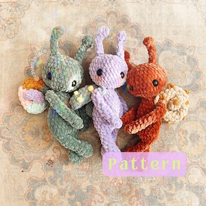 May include: Three crocheted amigurumi toys in the shape of bugs. The bugs are green, purple, and orange. The green bug has a pink and white flower on its back. The purple bug has a white flower on its back. The orange bug has a brown flower on its back. The toys are on a beige rug. The text "Pattern" is written in white letters on a pink background.