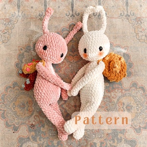 Könnte beinhalten: Zwei gehäkelte Hasen-Plüschtiere, eines rosa und eines weiß, mit einem braunen gehäkelten Rucksack auf dem weißen Hasen. Die Hasen halten sich an den Händen und liegen auf einem gemusterten Teppich. Der Text "Pattern" ist in Orange auf dem Teppich geschrieben.