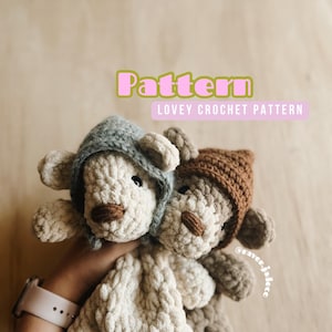 May include: Two crocheted teddy bear loveys with hooded hats. One teddy bear is beige with a gray hooded hat and the other teddy bear is brown with a brown hooded hat. The teddy bears are held in a hand with a white watch on the wrist.