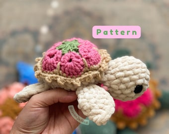May include: A crocheted turtle amigurumi toy with a pink and green shell. The turtle is made with a soft, plush yarn and has a sweet, friendly expression. The toy is being held in a hand with the Instagram handle "@ravenjadeco" visible.
