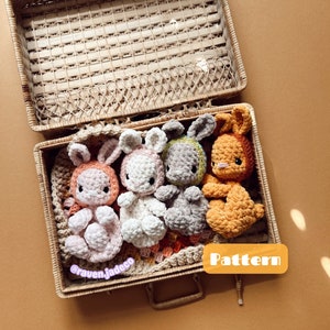 May include: Four crocheted bunny plushies in a wicker basket. The bunnies are in various colours, including pink, white, grey, and orange. The basket is lined with a crocheted blanket. The text "Pattern" is visible on a label in the lower right corner of the image.